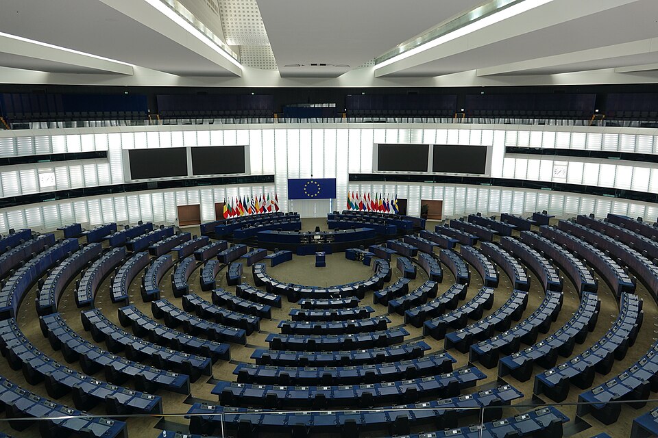 The empty European Parliament hemicycle in Strasbourg, with curved rows of seats and national flags.